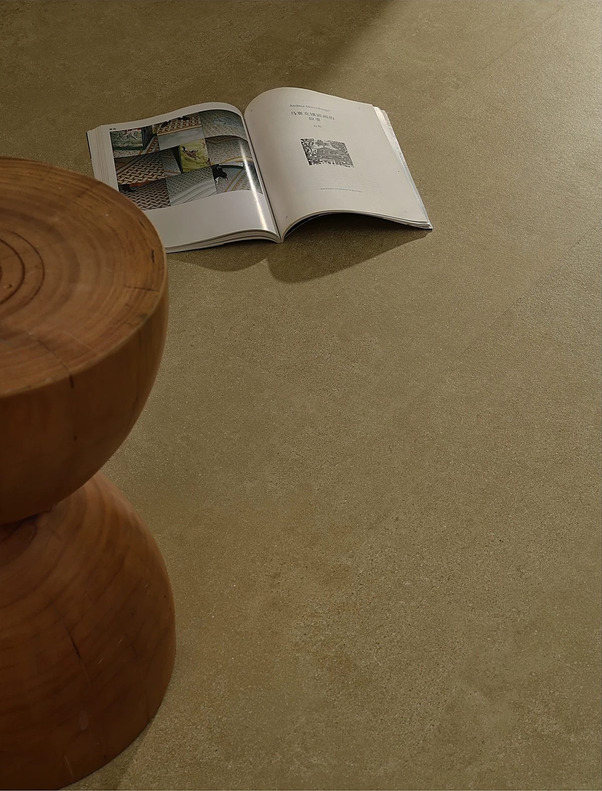 A close view of a sculpted timber side table and an open book on a green-grey stone-look floor