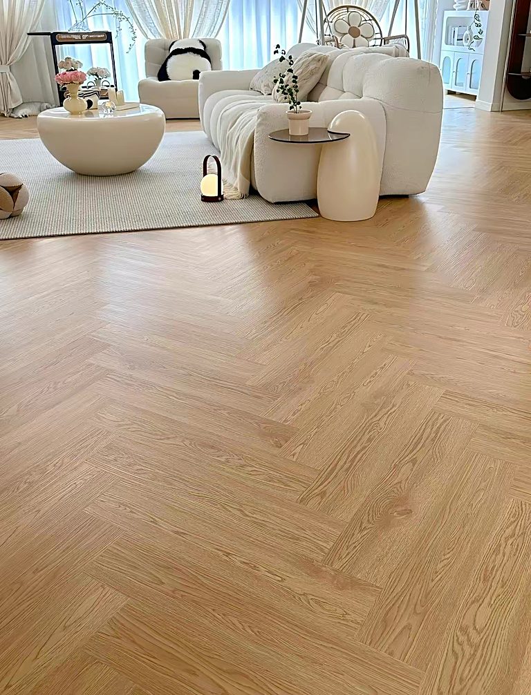 Herringbone-pattern flooring in a light oak finish laid across a bright living room, with a sofa and coffee table in the background.