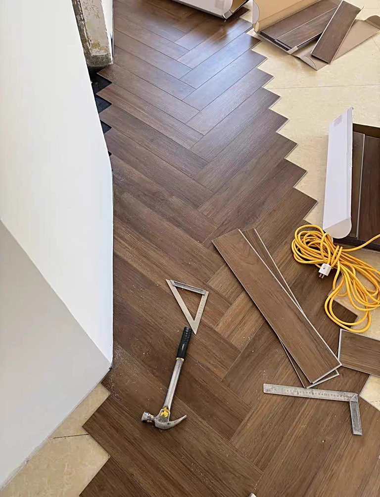 Dark wood-look herringbone planks during installation, with a hammer, set square and coiled cable resting on the floor.