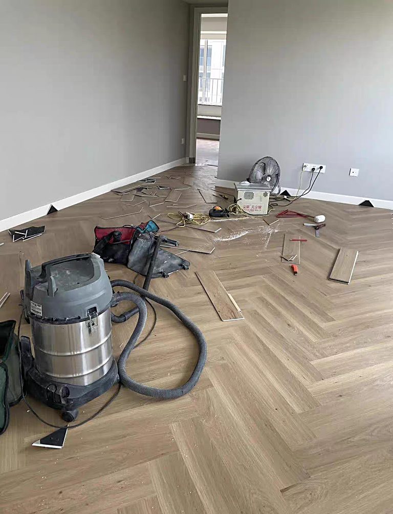 A room mid-installation with herringbone flooring partly laid, a vacuum cleaner, and installation tools on the floor.